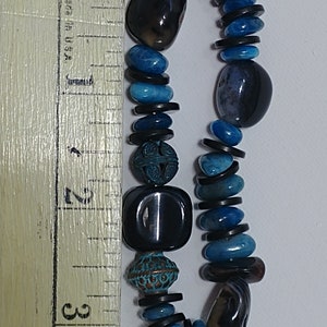 May include: A beaded bracelet featuring a mix of smooth black stones, vibrant blue polished stones, and two ornate, textured copper-toned metal beads. A wooden ruler is placed alongside the bracelet, showing a length of approximately 7.6 cm.