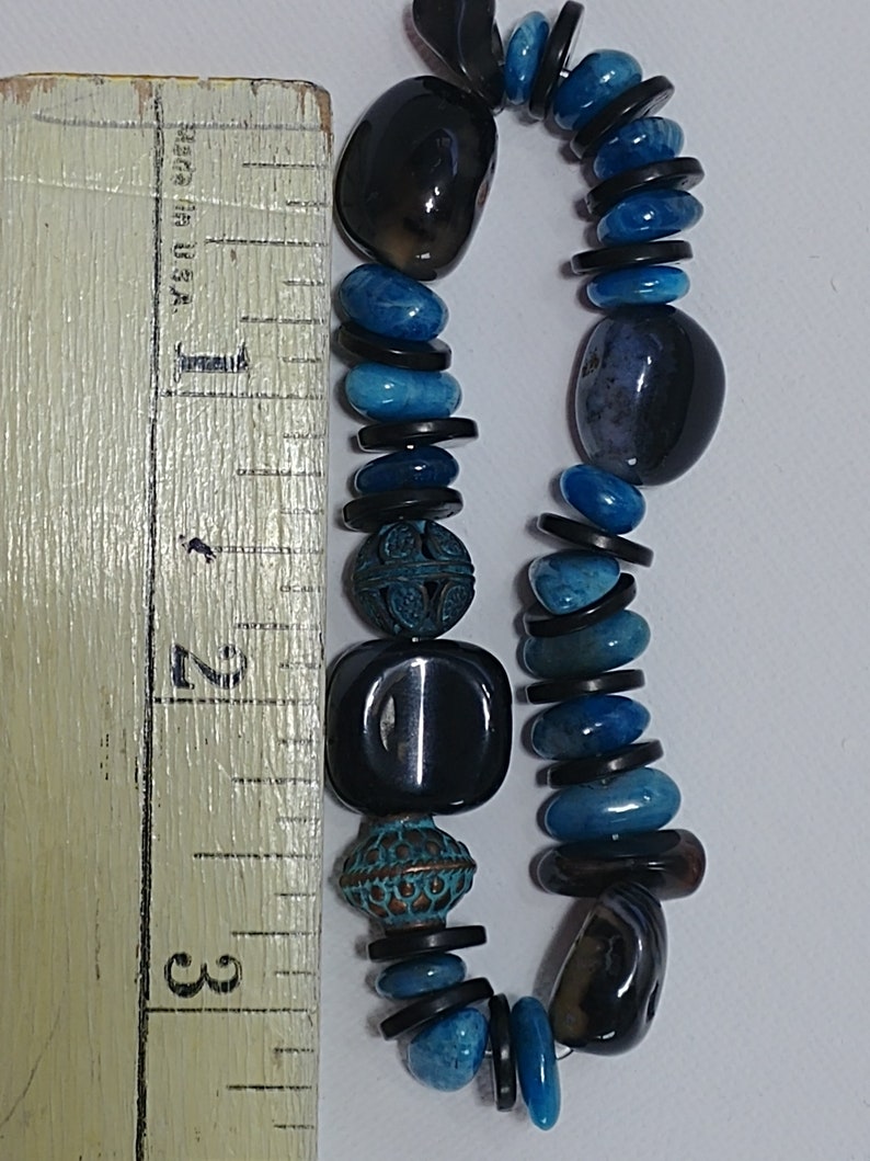 May include: A beaded bracelet featuring a mix of smooth black stones, vibrant blue polished stones, and two ornate, textured copper-toned metal beads. A wooden ruler is placed alongside the bracelet, showing a length of approximately 7.6 cm.