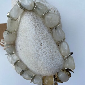 May include: A stretch bracelet composed of polished, translucent white stone beads interspersed with silver-toned metal spacers. One bead features an ornate, gold-toned filigree metal cap. The beads are strung on a visible, light-colored elastic cord.
