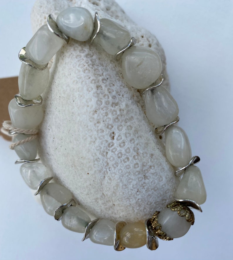 May include: A stretch bracelet composed of polished, translucent white stone beads interspersed with silver-toned metal spacers. One bead features an ornate, gold-toned filigree metal cap. The beads are strung on a visible, light-colored elastic cord.