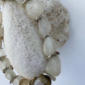 May include: A bracelet composed of smooth, off-white, irregular stone beads interspersed with thin, silver-colored metal spacers. One bead features a decorative, textured gold-toned metal cap at its base, adding a subtle metallic contrast.