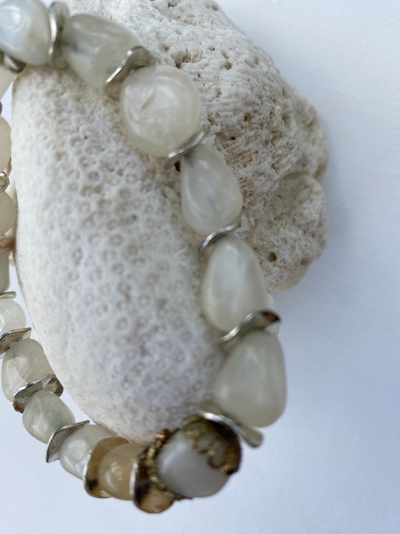 May include: A bracelet composed of smooth, off-white, irregular stone beads interspersed with thin, silver-colored metal spacers. One bead features a decorative, textured gold-toned metal cap at its base, adding a subtle metallic contrast.