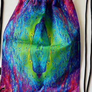 May include: A drawstring backpack featuring a vibrant, swirling pattern of iridescent colors including bright blue, lime green, magenta, and purple. The bag has a gathered top closure and thin black cord straps attached to metal grommets at the bottom.