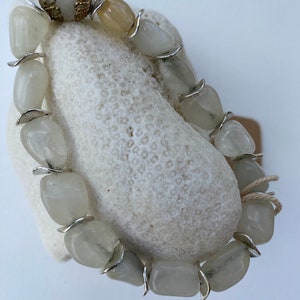 May include: A bracelet composed of polished, translucent off-white stone beads separated by curved silver-toned metal spacers. One end of the bracelet features a decorative gold-toned filigree bead cap, and a thin beige string is tied near the clasp.