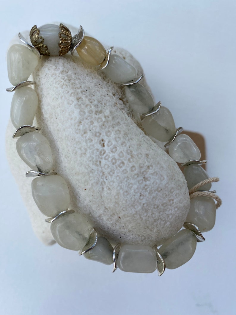 May include: A bracelet composed of polished, translucent off-white stone beads separated by curved silver-toned metal spacers. One end of the bracelet features a decorative gold-toned filigree bead cap, and a thin beige string is tied near the clasp.