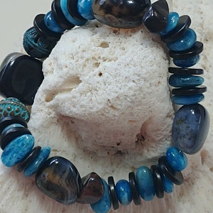 May include: A stretch bracelet featuring a mix of polished blue stone beads, smooth black disc-shaped spacers, and larger dark brown and grey patterned stones. The design includes two textured, round, teal-coloured metal beads with intricate patterns.