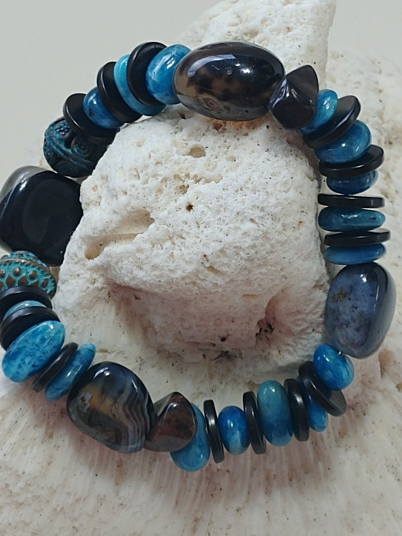 May include: A stretch bracelet featuring a mix of polished blue stone beads, smooth black disc-shaped spacers, and larger dark brown and grey patterned stones. The design includes two textured, round, teal-coloured metal beads with intricate patterns.