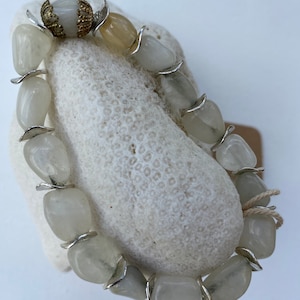 May include: A bracelet composed of smooth, translucent, pale gray tumbled stones separated by small, curved silver-toned metal spacers. One end of the bracelet features a decorative, filigree-style gold-toned metal cap accenting a single stone.