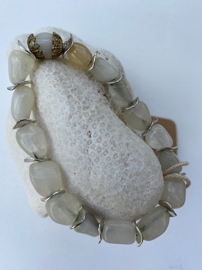 May include: A bracelet composed of smooth, translucent, pale gray tumbled stones separated by small, curved silver-toned metal spacers. One end of the bracelet features a decorative, filigree-style gold-toned metal cap accenting a single stone.