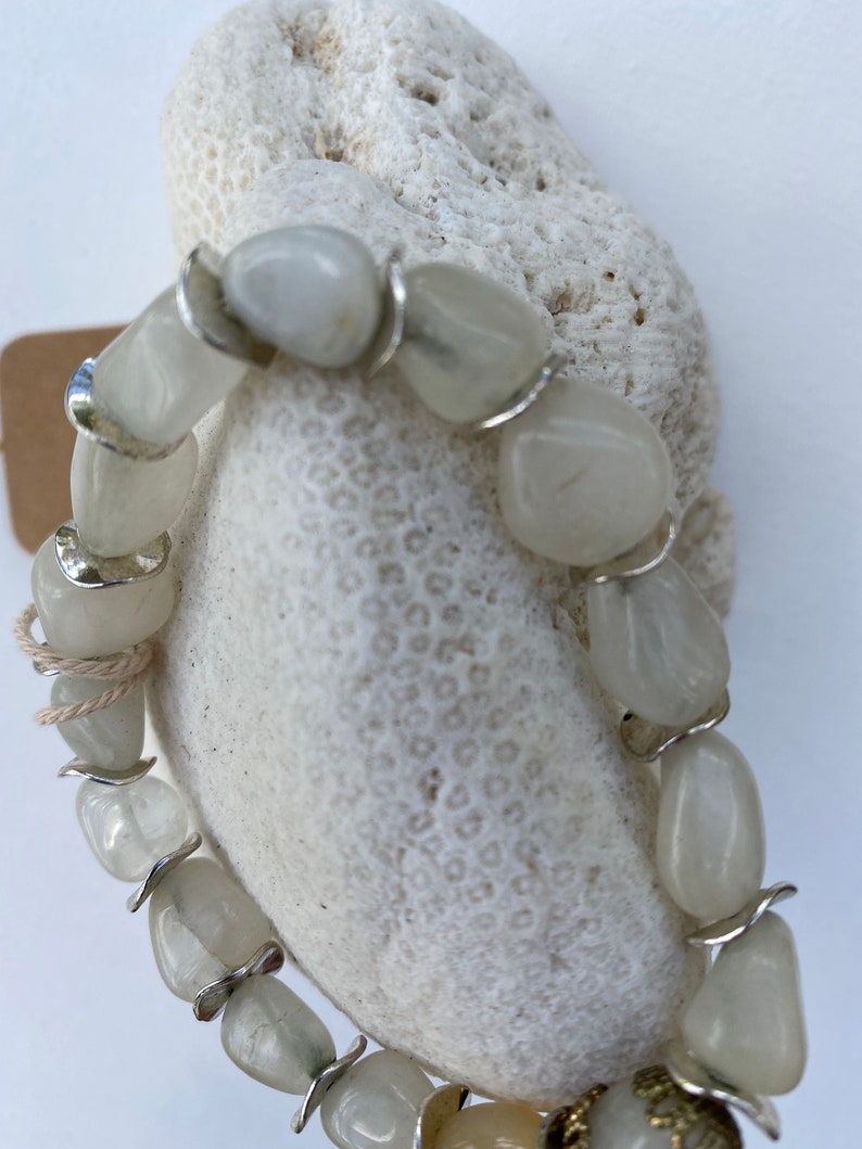 May include: A bracelet composed of smooth, translucent white tumbled stones alternating with curved silver-toned metal spacers. The stones have a polished, rounded appearance, and the metal components provide a reflective contrast to the pale, milky texture of the stones.