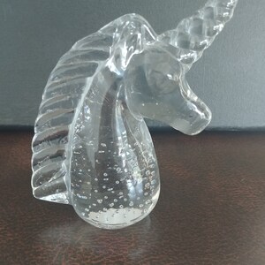 May include: A clear glass unicorn head figurine featuring a textured mane and a spiraled horn. The base of the sculpture contains small, trapped air bubbles within the glass, creating a distinct visual effect throughout the lower portion.