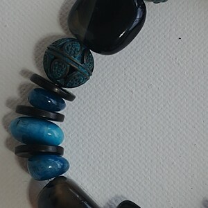 May include: A beaded necklace featuring a sequence of smooth black polished stones, textured copper-toned beads with a teal patina finish, and vibrant blue oval-shaped stones, all separated by thin, flat, circular black spacers.