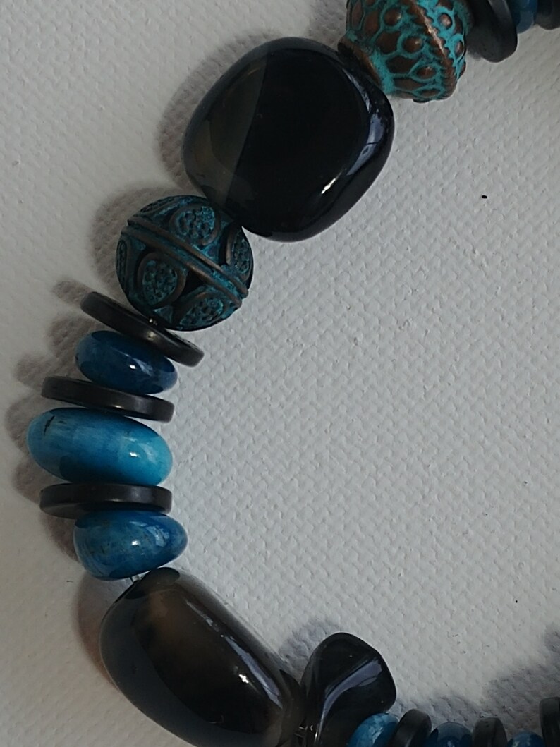 May include: A beaded necklace featuring a sequence of smooth black polished stones, textured copper-toned beads with a teal patina finish, and vibrant blue oval-shaped stones, all separated by thin, flat, circular black spacers.