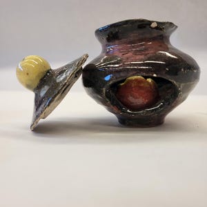 Puede incluir: Una pequeña olla de cerámica negra y morada con tapa. La olla tiene un agujero en el lateral y un objeto rojo y amarillo en su interior. La tapa tiene un pomo amarillo.