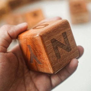 May include: A wooden cube with the letters "Y", "O", and "N" etched on its sides. The cube is held in a hand, with other wooden blocks visible in the background. The wood has a warm, reddish-brown tone.