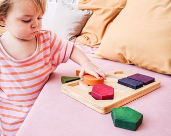 May include: A child in a pink and orange striped shirt plays with a wooden puzzle on a pink surface. The puzzle has various colored geometric shapes, including a green hexagon, and an orange circle. The background features pillows and a white patterned wall.