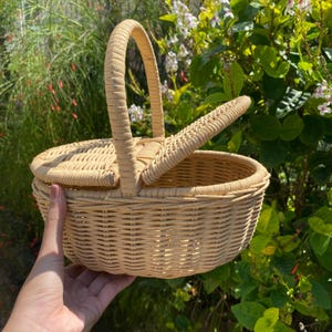 Op de afbeelding: Een lichtbruine picknickmand van riet met een handvat en een scharnierend deksel. De mand is rond met een geweven ontwerp en wordt voor een groene, bladerrijke achtergrond gehouden. Het deksel is gedeeltelijk open.