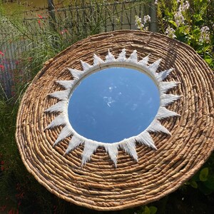 May include: Circular woven wall mirror in natural brown, featuring a white sunburst design around the edge. The mirror reflects the sky. The mirror is surrounded by green foliage.