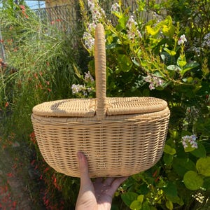 Op de afbeelding: Een lichtbruine geweven picknickmand met een handvat en een deksel. De mand wordt door een hand vastgehouden tegen een achtergrond van groen gebladerte en bloemen. De mand is gemaakt van geweven riet.