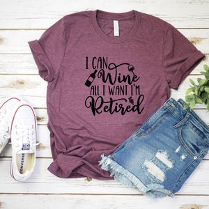 May include: A maroon t-shirt with the text "I can wine all I want I'm retired" in black lettering. The shirt is displayed on a white wooden surface with a pair of white sneakers and blue denim shorts.