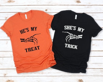 May include: Two t-shirts with a skeleton hand graphic and text. The orange shirt says "He's my treat" and the black shirt says "She's my trick".
