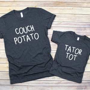 May include: Two dark gray t-shirts with white text. The larger shirt says "Couch Potato" and the smaller shirt says "Tator Tot".