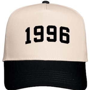 May include: A baseball cap with a cream-colored crown and a black brim. The number "1996" is printed in bold black letters across the front of the cap. The cap has a black button on top.