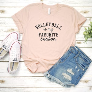 May include: A light pink t-shirt with the text "VOLLEYBALL is my FAVORITE season" in black. The shirt is paired with denim shorts and white sneakers with red accents. The items are arranged on a white wooden surface.