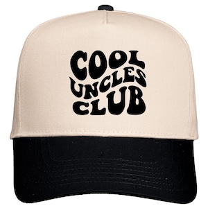 May include: A baseball cap with a cream-colored crown and a black brim. The front of the cap features the words "COOL UNCLES CLUB" in large, black, retro-style lettering. The cap has a curved brim and a black button on top.