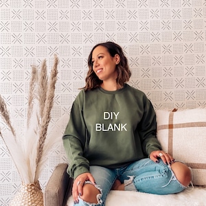 May include: An olive green sweatshirt with the text "DIY BLANK" in white block letters. The sweatshirt is worn with light blue ripped jeans. The background features a patterned wallpaper and decorative elements.