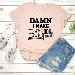 以下が含まれることがあります： ピンクのTシャツに黒い文字で「DAMN i MAKE 50 LOOK good」と書かれています。Tシャツは白い木製の表面に置かれ、白いスニーカーと青いデニムのショートパンツが置かれています。
