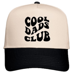 May include: A baseball cap with a cream-colored crown and a black brim. The front of the cap features the words "COOL DADS CLUB" in large, bold, black letters. The cap has a curved brim and a button on top.
