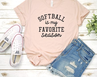 May include: A pink t-shirt with the text "Softball is my favorite season" in black lettering. The shirt is laid out on a white wooden surface with a pair of white sneakers and a pair of blue denim shorts.