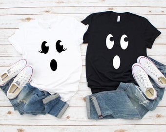 May include: Two t-shirts, one white and one black, each with a ghost face design. The white shirt has a ghost face with eyelashes, while the black shirt has a simpler design. Both are paired with blue jeans and white trainers.