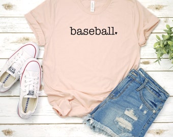May include: A pink t-shirt with the word "baseball" printed on it in black. The shirt is laid out on a white wooden surface with a pair of white sneakers and a pair of blue denim shorts.