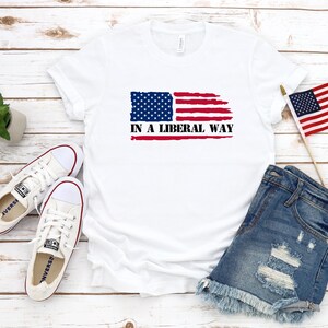 以下が含まれることがあります： かすれたアメリカ国旗のグラフィックと黒い文字で「IN A LIBERAL WAY」とプリントされた白い半袖Tシャツ。白いキャンバス地のスニーカー、小さな多肉植物、ミニチュアのアメリカ国旗が一緒に配置されています。