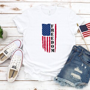 以下が含まれることがあります： 白いTシャツに、くたびれたアメリカ国旗のデザインと、縦に「FREEDOM」という文字がプリントされています。Tシャツは、白い木製の表面に置かれ、白いキャンバス地の靴、小さな鉢植えの植物、フリンジの付いたデニムのショートパンツが置かれています。