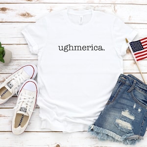 以下が含まれることがあります： 前面に黒い文字でughmerica.とプリントされた白い半袖Tシャツ。白いキャンバス地のスニーカー、ダメージ加工が施された青いデニムのショートパンツ、木製の棒についた小さなアメリカ国旗が添えられています。