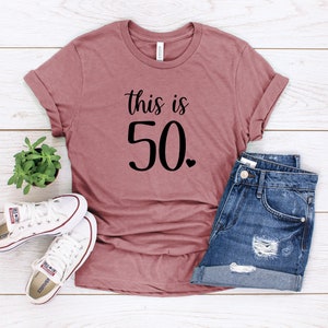 May include: A mauve t-shirt with the text "This is 50." in black lettering. The shirt is laid flat on a white wooden surface with a pair of white sneakers and denim shorts.