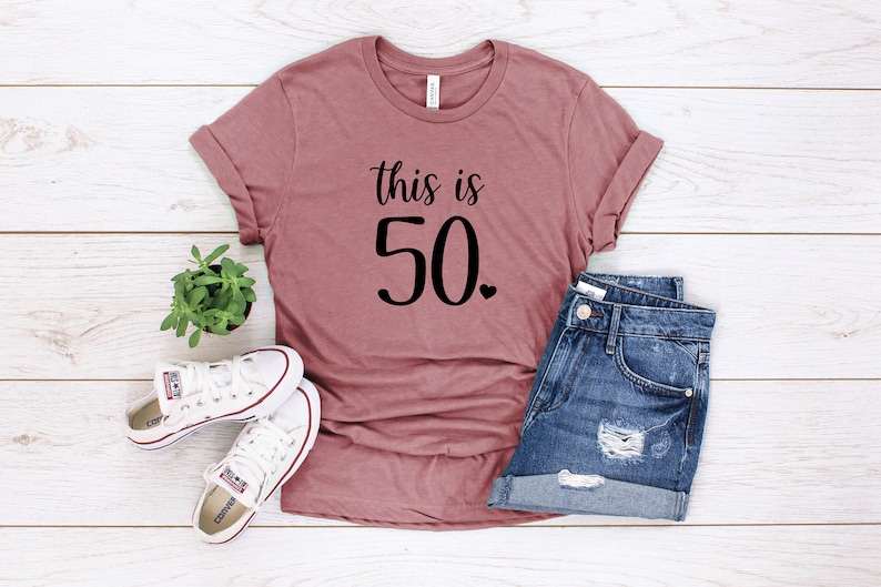 May include: A mauve t-shirt with the text "This is 50." in black lettering. The shirt is laid flat on a white wooden surface with a pair of white sneakers and denim shorts.