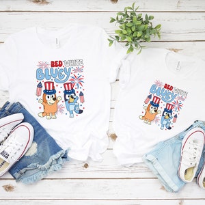 以下が含まれることがあります： 「RED WHITE BLUEY」の文字と、愛国的な帽子をかぶった漫画のキャラクターが描かれた2枚の白いTシャツ。デニムショーツと白いスニーカーと組み合わせて、カジュアルで祝祭的なテーマを表現しています。