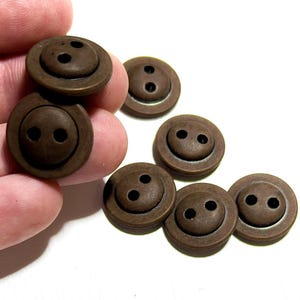 May include: A collection of brown, round buttons with two holes each. The buttons have a slightly raised outer ring and a darker brown color in the center. The buttons are scattered on a white surface.