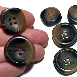 May include: A collection of eight brown and black marbled buttons with four holes each. Two buttons are held in a hand, while the others are scattered on a white surface. The buttons have a slightly rounded, square shape.