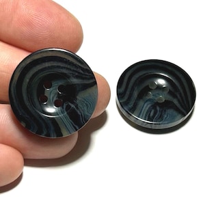 May include: Two round buttons with a marbled design in shades of black, gray, and blue. Each button has four holes for sewing. One button is held by a hand, while the other rests on a white surface.