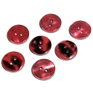 Op de afbeelding: Zeven rode knopen met elk twee gaten liggen verspreid op een witte ondergrond. De knopen hebben een gemarmerd uiterlijk, met donkerdere rode en zwarte wervelingen. De knopen zijn rond en lijken van een glanzend materiaal te zijn gemaakt.