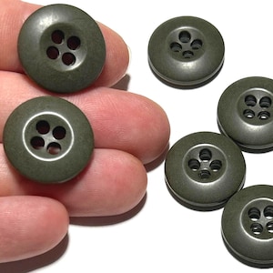 May include: A collection of olive green buttons with four holes each. The buttons are round and have a slightly raised edge. Some buttons are held in a hand, while others are scattered on a white surface. These buttons are suitable for sewing and craft projects.