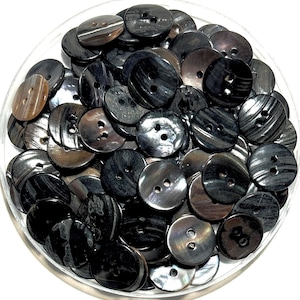 May include: A clear container filled with a collection of black and gray buttons. The buttons have two holes and a slightly iridescent sheen. The buttons are round and vary in size and pattern.