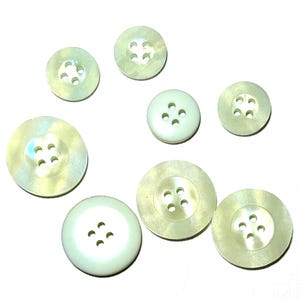 May include: A collection of eight off-white buttons of varying sizes. Each button has four holes and a slightly iridescent sheen. The buttons are arranged on a white surface, showcasing their circular shape and subtle color variations.