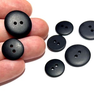May include: A collection of black, round buttons with two holes each. The buttons vary in size and are displayed against a white background. Some buttons are held in a hand, showcasing their texture and color.