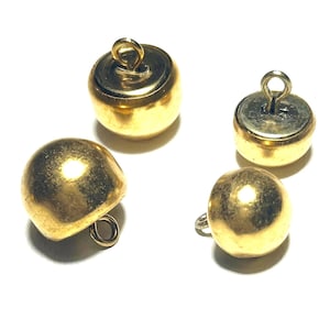 Może przedstawiać: Cztery, złote, zabytkowe guziki o zaokrąglonym, kulistym kształcie. Każdy guzik ma małą pętelkę na górze do przyszywania. Guziki znajdują się na białym tle, a światło odbija się od błyszczącej powierzchni.